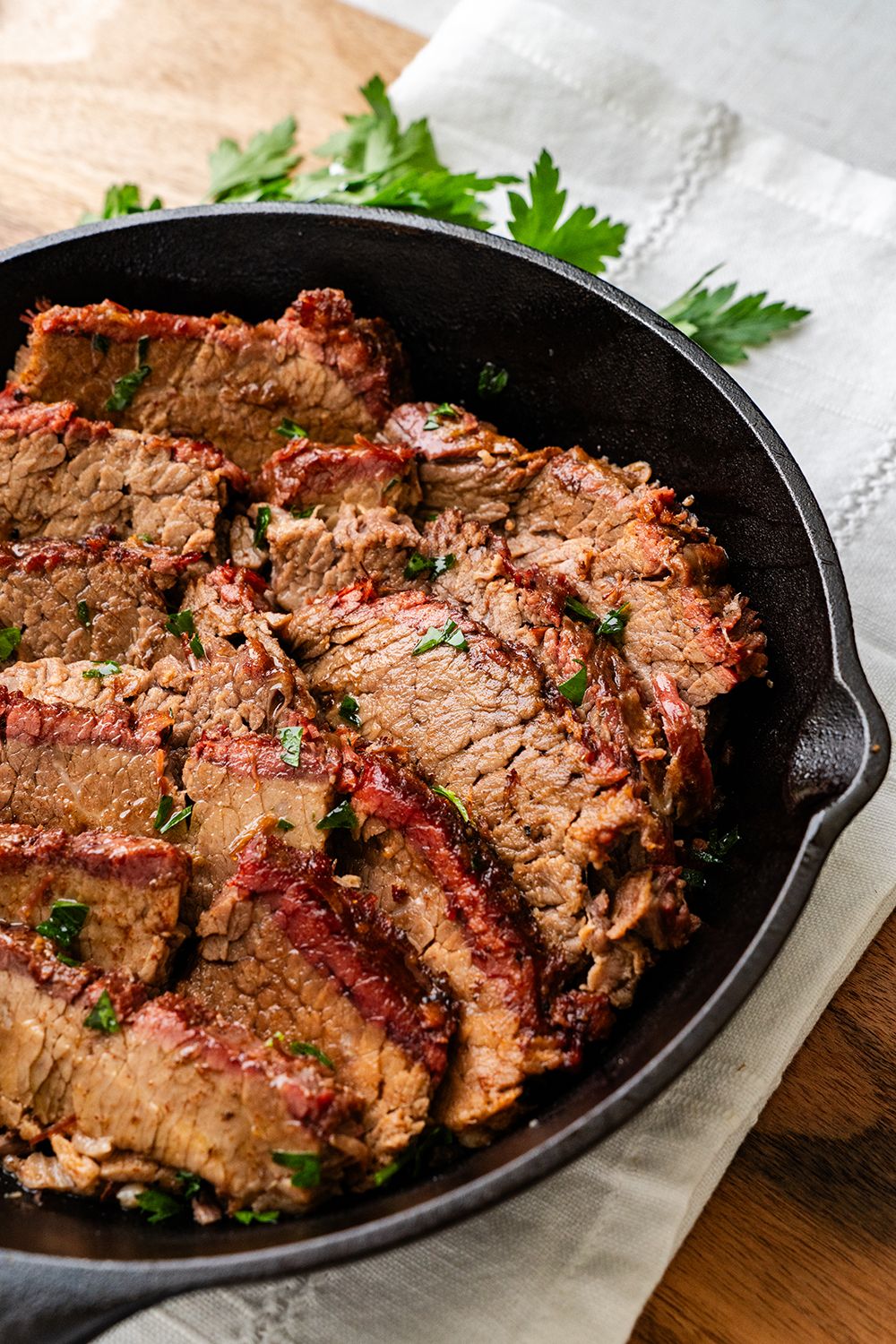 Professionally lit photo of steak slices in a cast-iron pan on linen with parsley garnishing the meat.