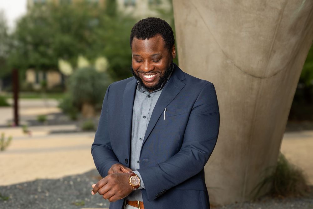 Professional man in navy blazer smiling outdoors near a tree in a well-landscaped setting.