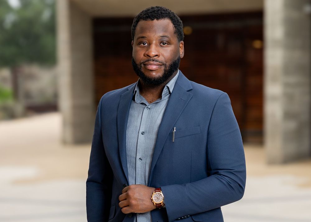 Professional Black man in navy blue blazer and dress shirt poses confidently outdoors near modern building.