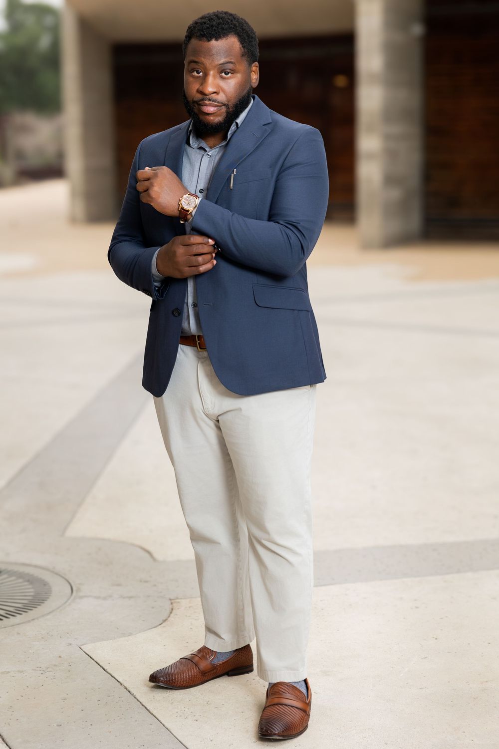 A well-dressed Black man in a navy blazer, grey pants, and brown loafers adjusts his cuff outdoors.