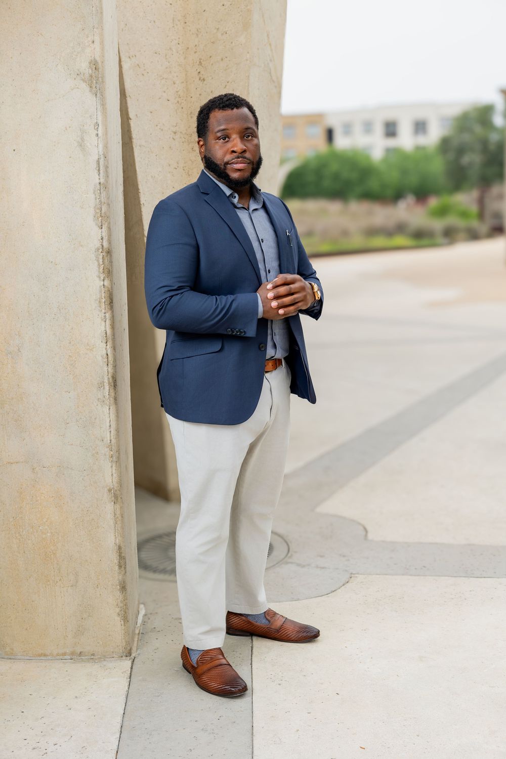 A well-dressed Black man in a navy blazer, white pants, and brown loafers poses outdoors near a stone column.