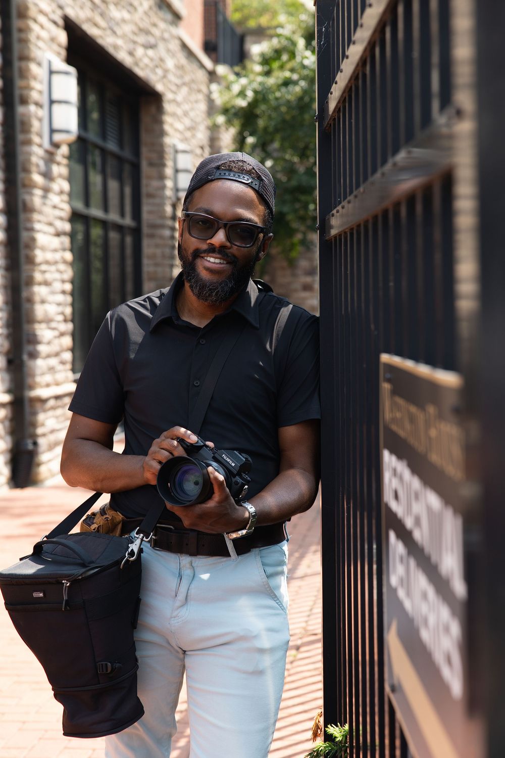 Smiling Black man with beard holding DSLR camera outdoors, wearing sunglasses and black shirt, leaning against dark metal gate.