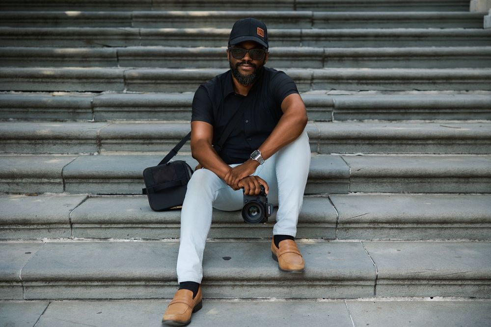 A smiling man in a black polo and cap sits on stone steps with a camera and bag on a sunny day.