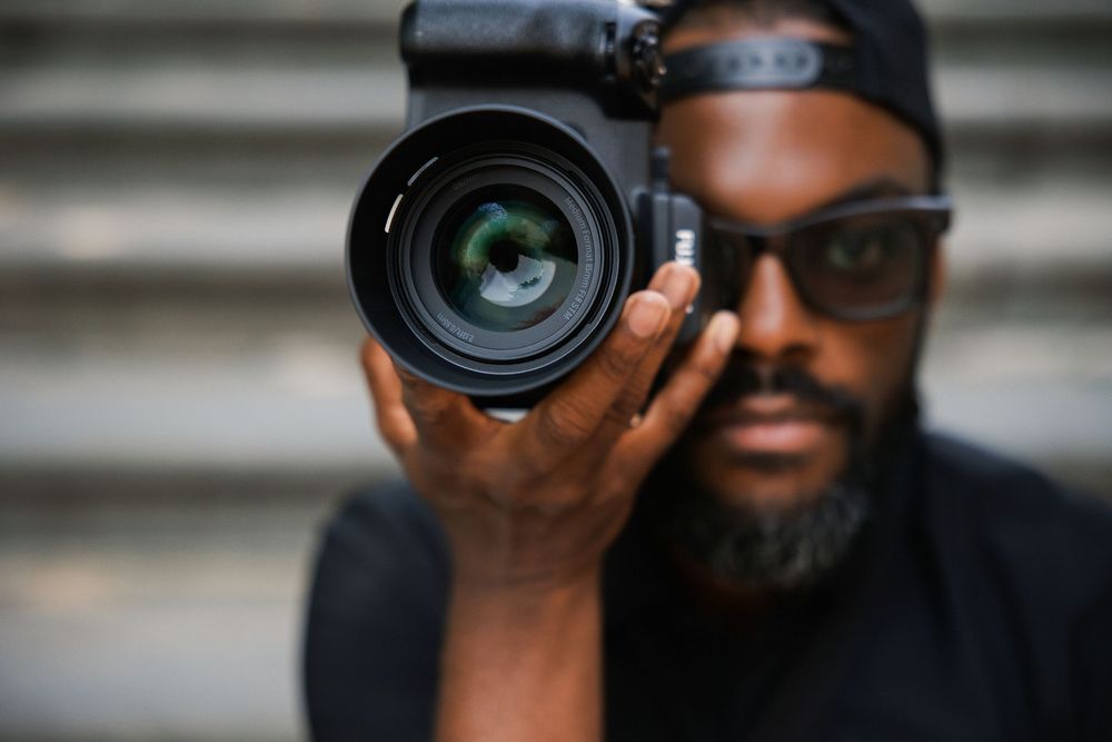 A Black male photographer holds a DSLR camera up to his eye, with a large lens in sharp focus in the foreground.