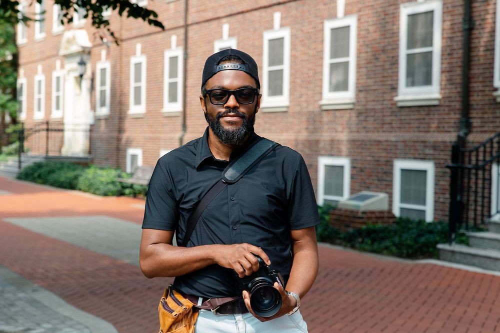 A stylish Black man wearing sunglasses and a black polo shirt holds a camera on a brick university campus path.