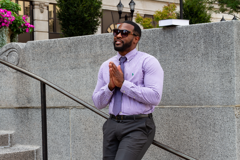Man in business attire sits thoughtfully on outdoor concrete steps among flowering plants.  L Visions Photography. Business professional