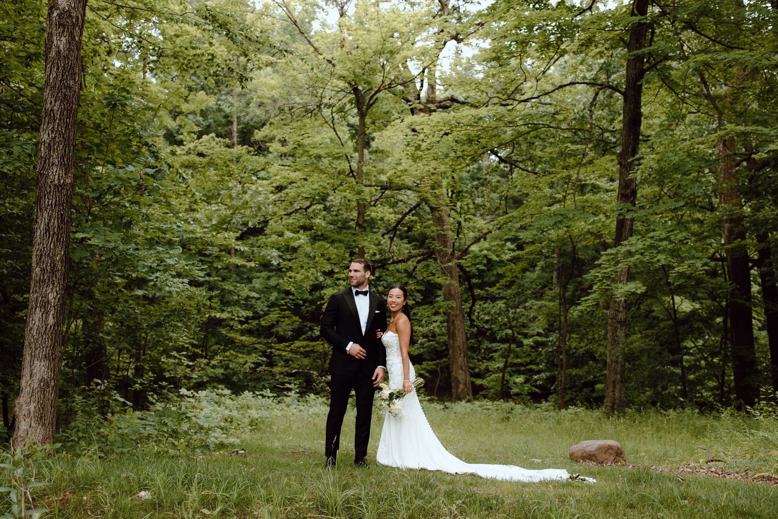 A couple in formal wedding attire walks through a lush green forest setting during their outdoor ceremony.