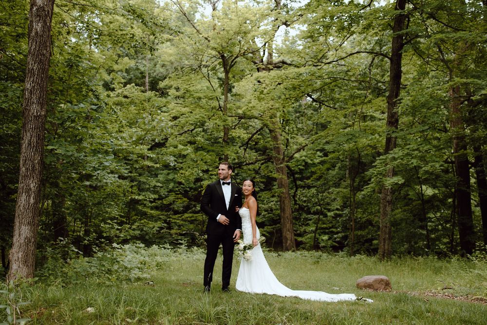A couple in formal wedding attire walks through a lush green forest setting during their outdoor ceremony.