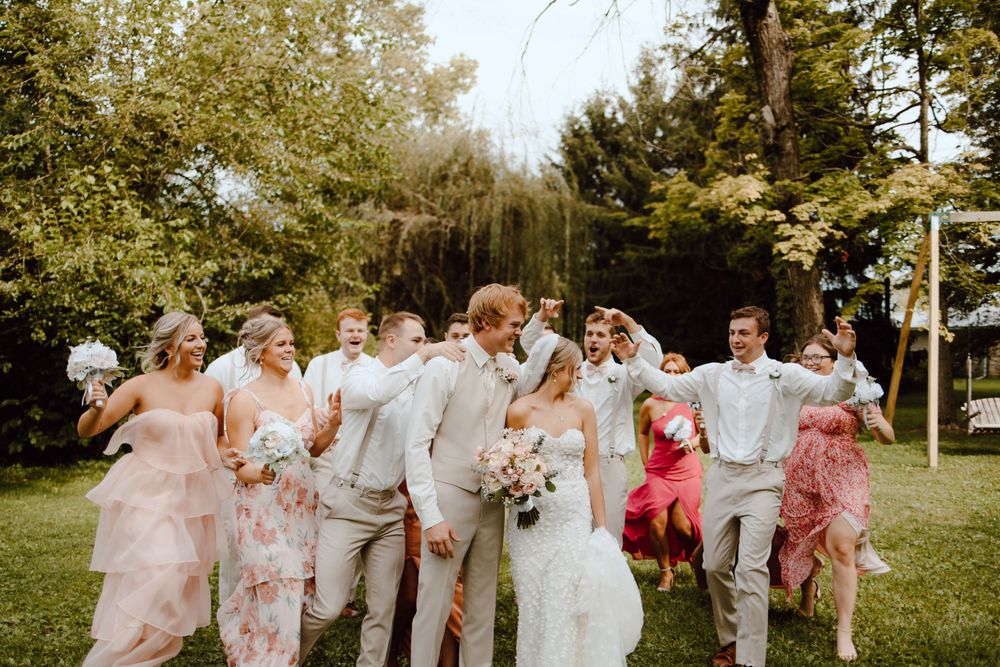 Wedding party wearing soft pink and gray outfits laughs together on a sunny day in a garden setting.