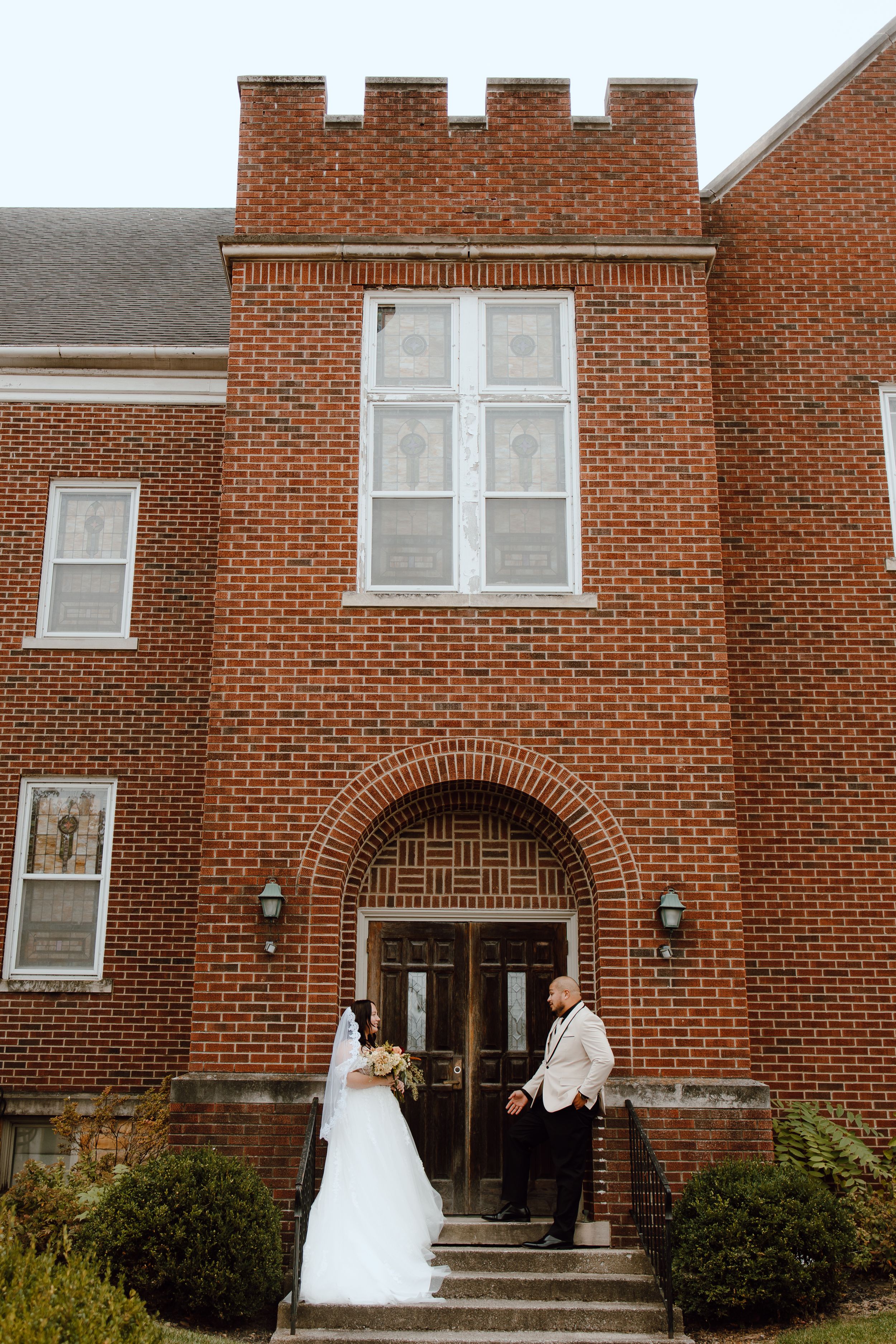 A bride in a white wedding dress poses in front of a traditional red brick church building entrance.