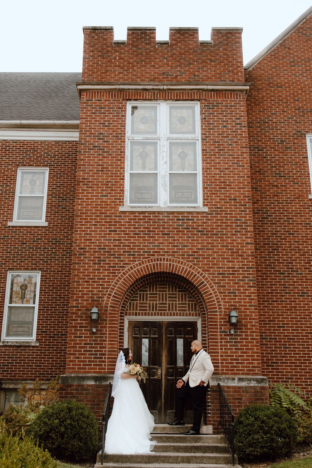 A bride in a white wedding dress poses in front of a traditional red brick church building entrance.