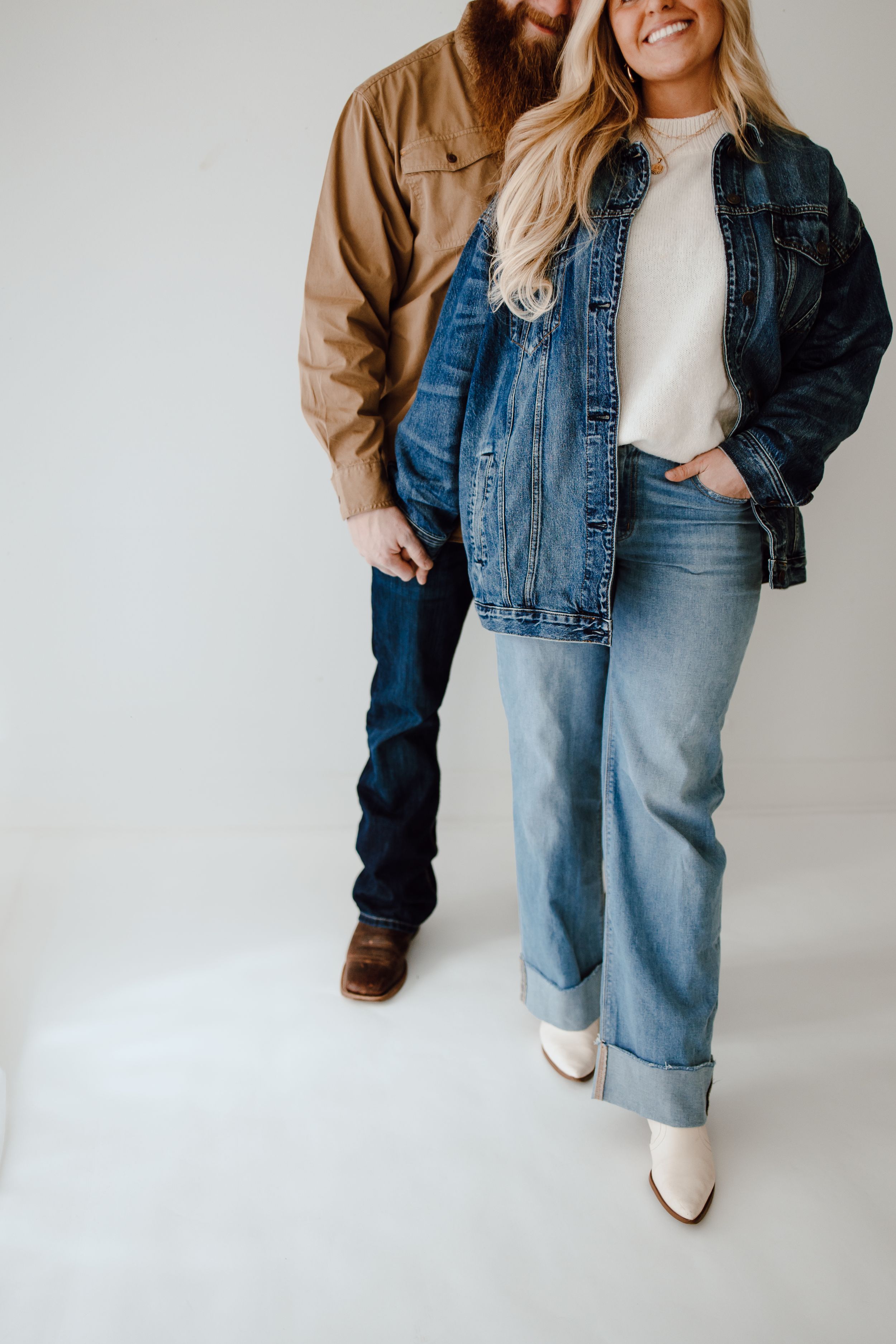 Couple wearing coordinated denim and neutral outfits with white boots and brown shoes on white background.
