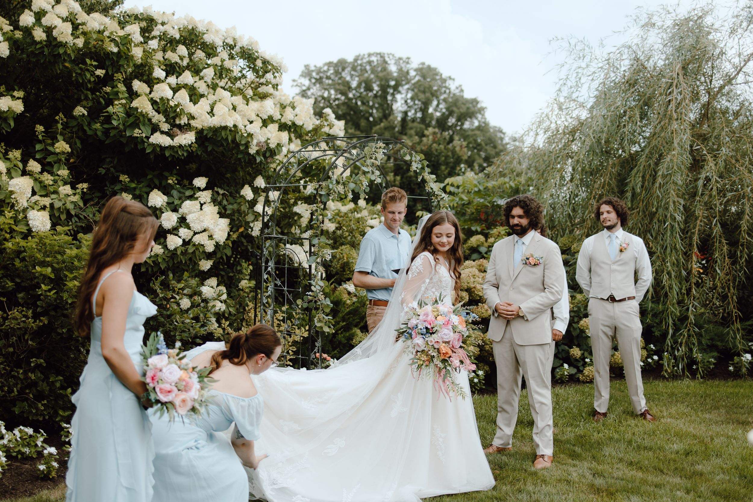 A wedding party stands in a garden setting with white roses and willow trees during an outdoor summer ceremony.