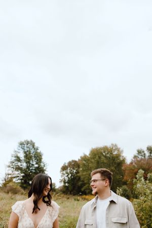 A couple stands together in a grassy field on an overcast autumn day with trees in the background.