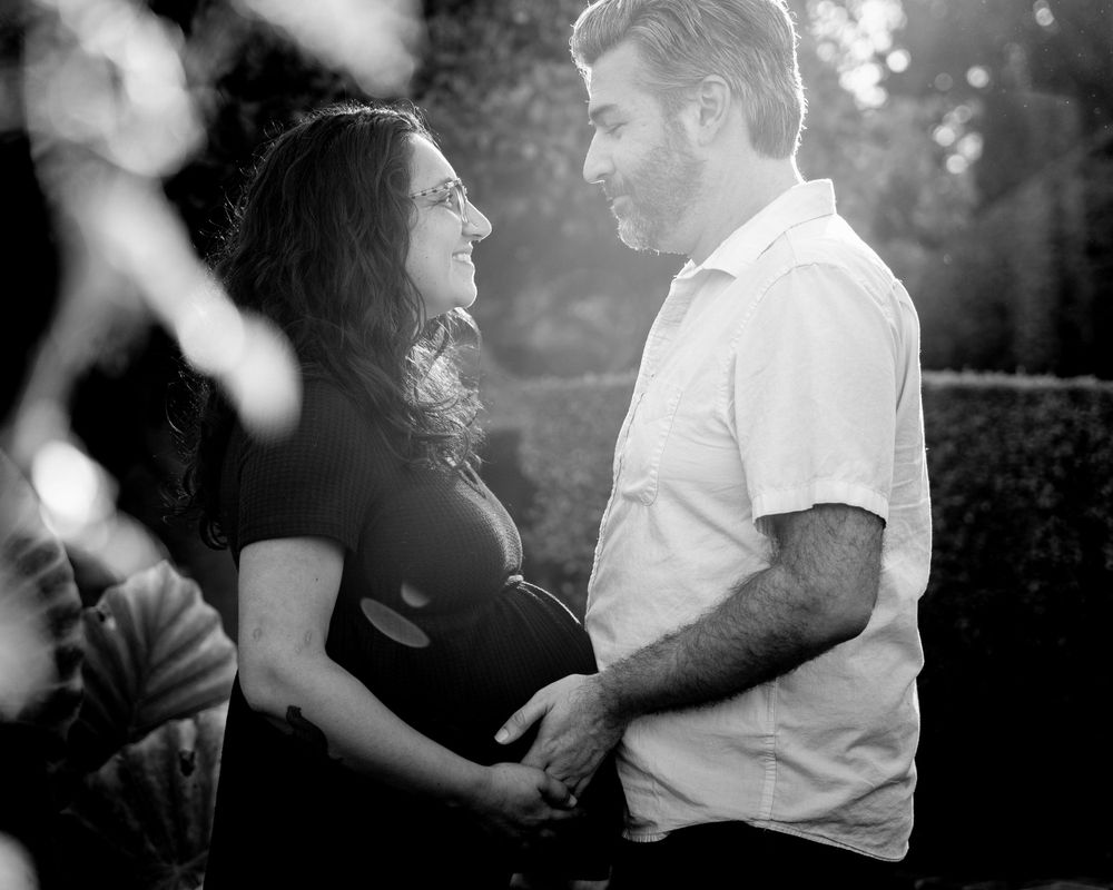 A black and white maternity photo shows a couple embracing outdoors in soft sunlight with bokeh effects.