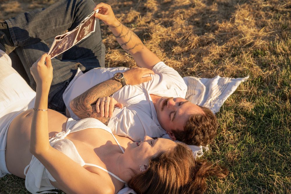 Couple lying on grass looking at an ultrasound photo together in warm golden sunlight.