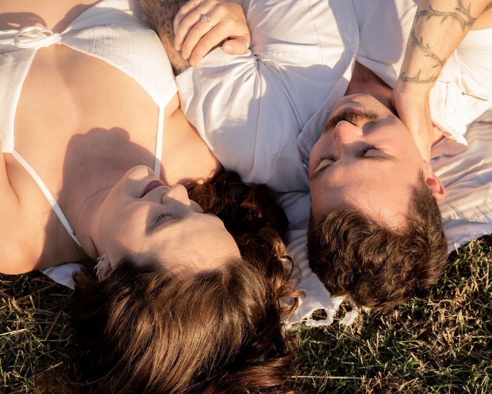 Couple lying on grass in white outfits, heads together, eyes closed, enjoying a peaceful romantic outdoor moment.