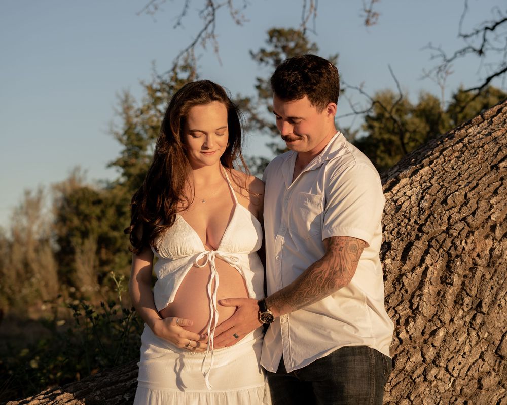 Couple sharing a tender maternity moment outdoors at golden hour, woman in white dress cradling baby bump.