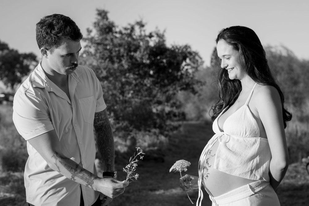 Black and white maternity photo of pregnant woman and man outdoors, sharing a tender moment with wildflowers.