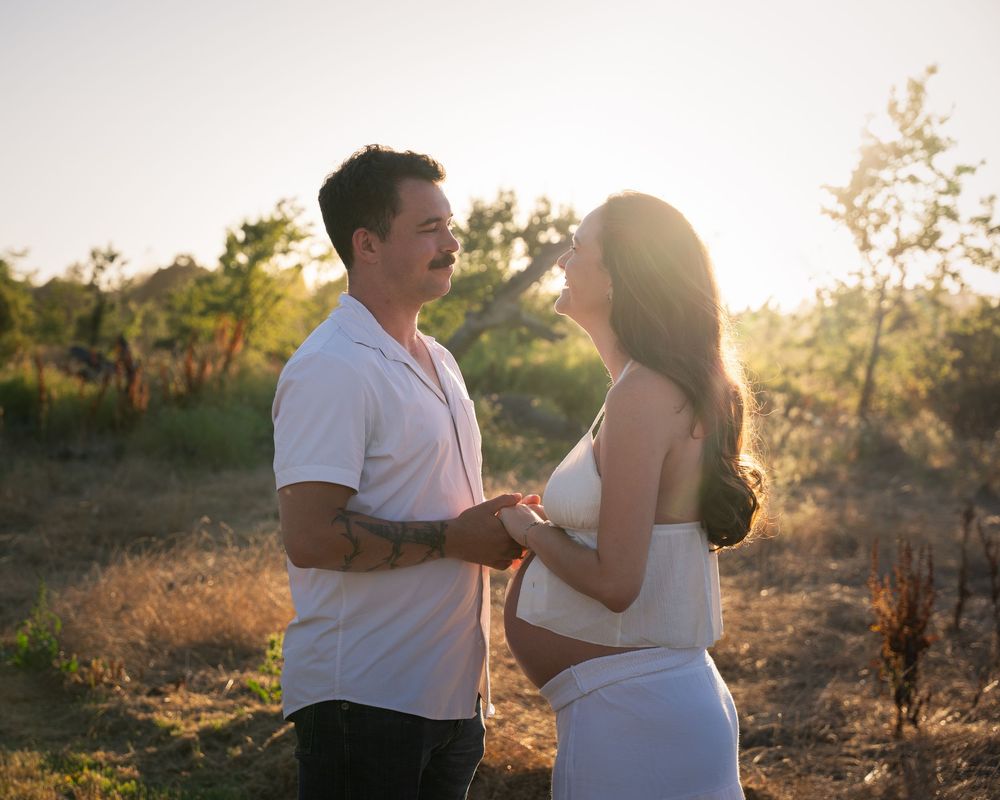 A couple holds hands in a sunlit field, the woman visibly pregnant, sharing a tender moment outdoors.