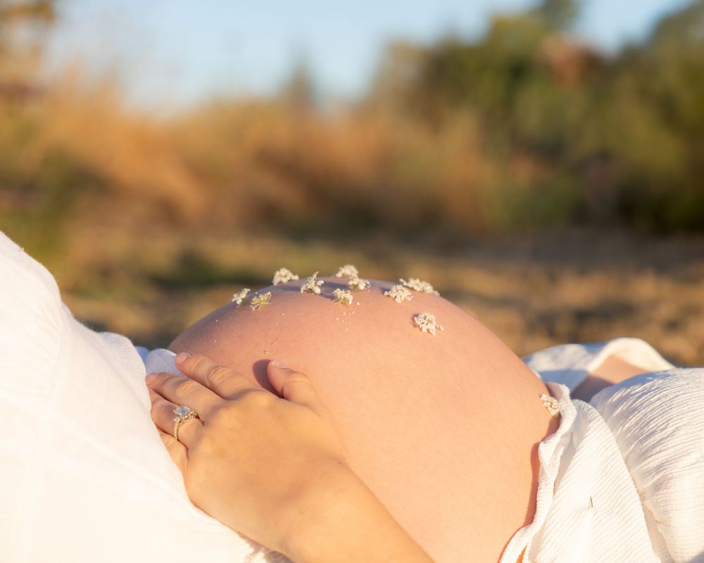 Close-up of a pregnant woman cradling her bare baby bump with small white flowers outdoors in golden sunlight.
