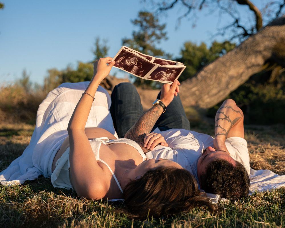 Couple lying on grass looking at ultrasound photo outdoors, expecting parents sharing pregnancy announcement moment.