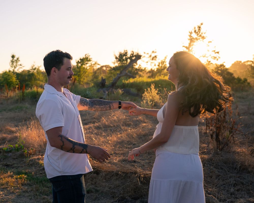 Couple holding hands at golden hour sunset in a dry field, man in white shirt with tattoos, woman in white dress.