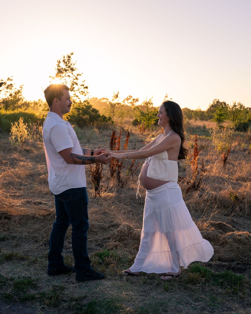 Couple holding hands in golden sunset field, pregnant woman in white dress showing baby bump.