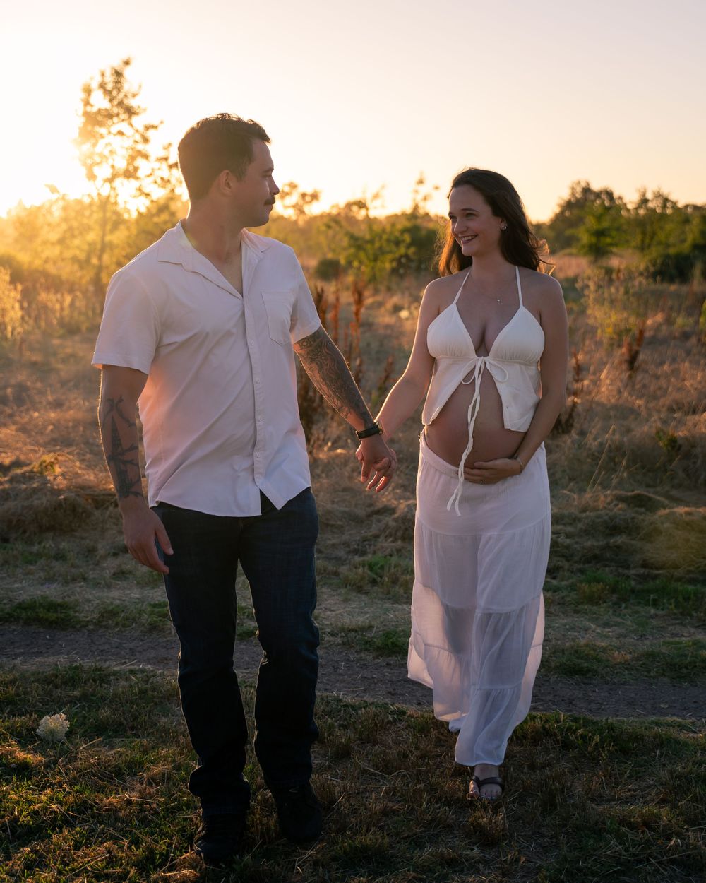 Couple in white outfits holding hands at golden hour, pregnant woman smiling in flowing dress outdoors.