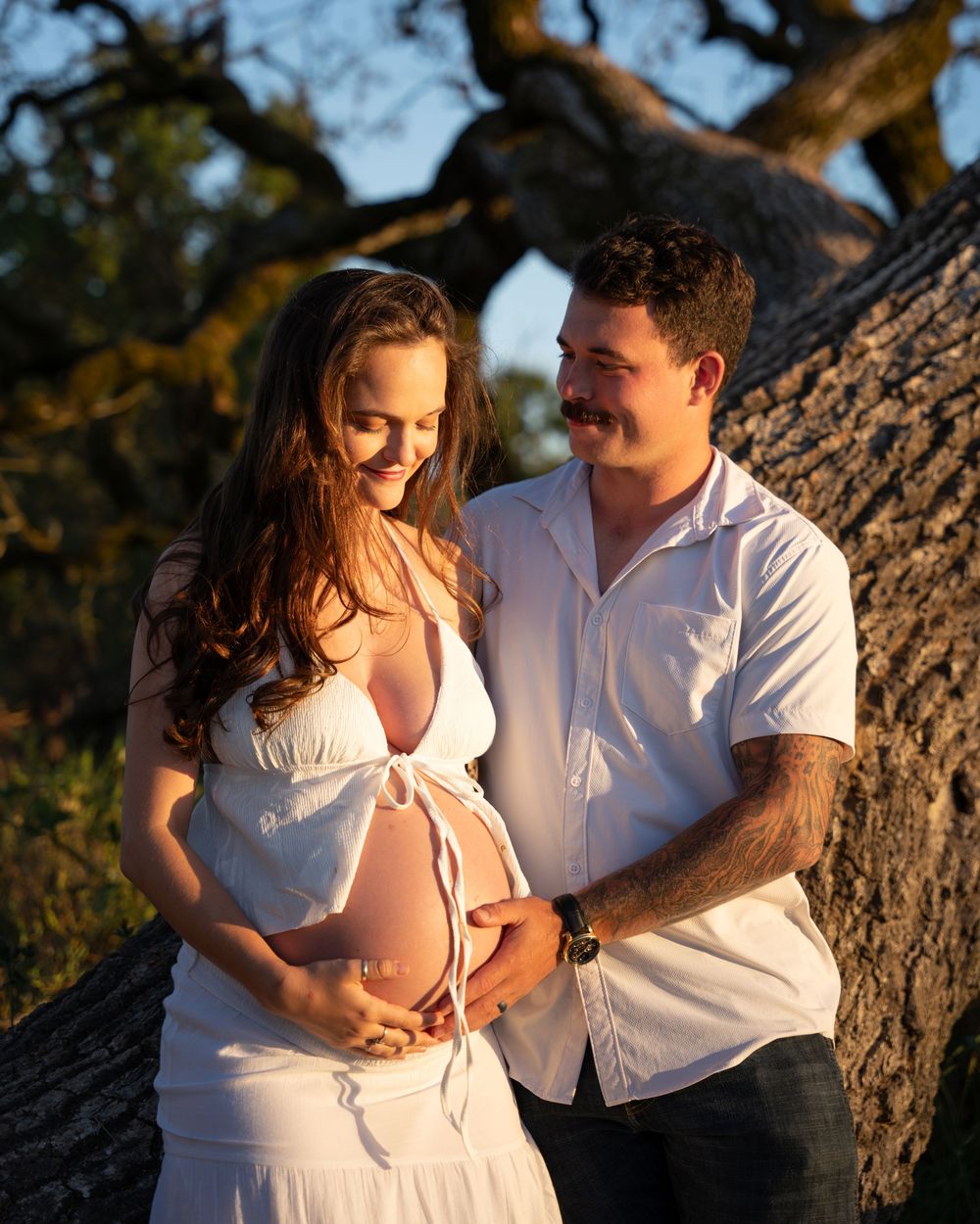 Couple cradling pregnant belly outdoors at golden hour near a large oak tree.