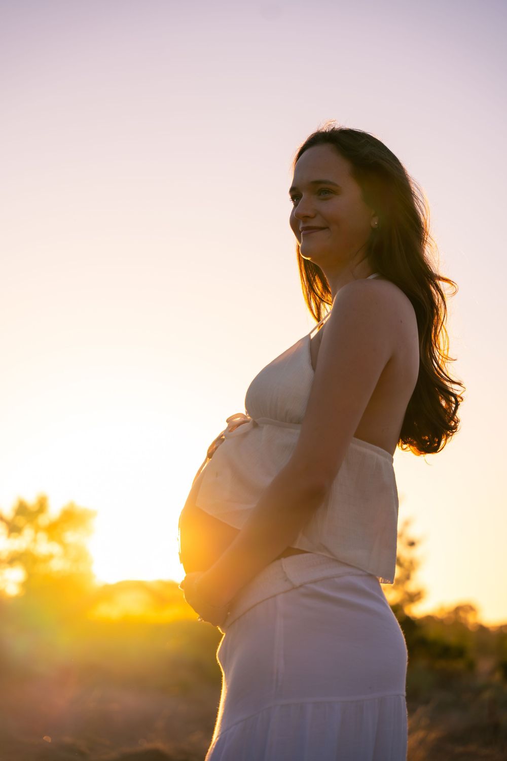 Pregnant woman in white dress holding belly at sunset with warm golden backlight.