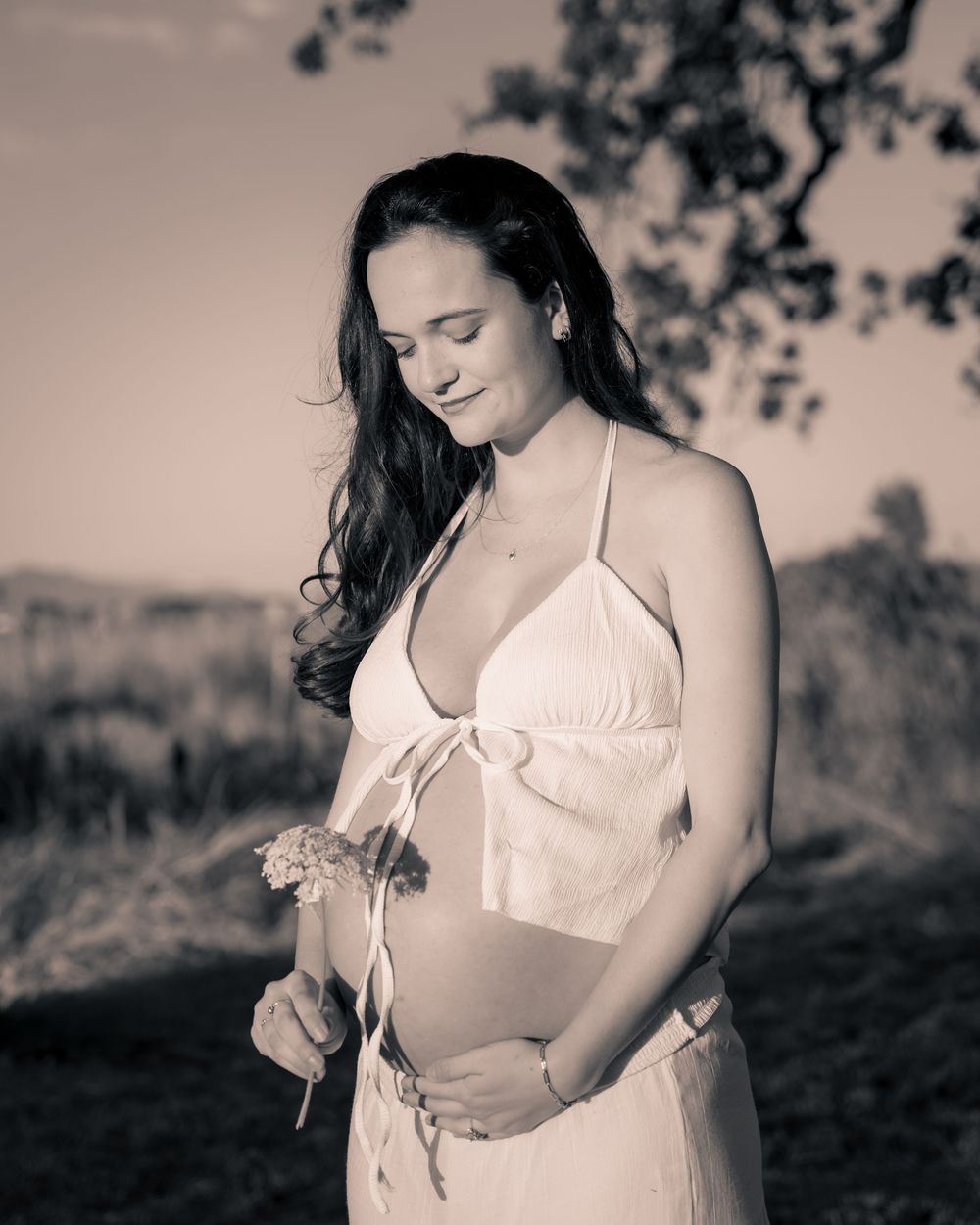 Pregnant woman in white halter dress holding flowers outdoors in a serene black and white maternity portrait.