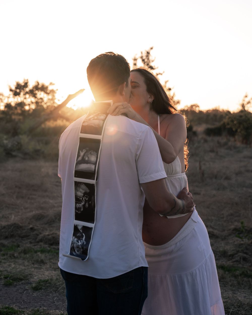 Couple sharing kiss at sunset, man holding ultrasound photos on his back during maternity shoot.