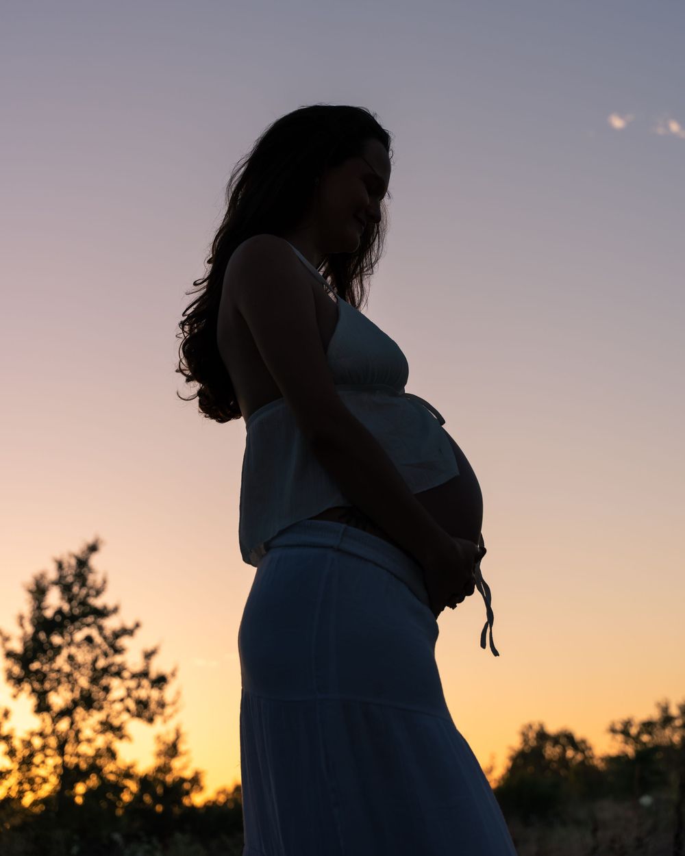 Silhouette of pregnant woman cradling baby bump against vibrant sunset sky in white dress.
