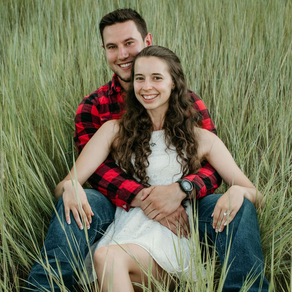 A couple poses together in tall grass wearing coordinated red plaid and white outfits.
engagement, surprise proposal, Montana