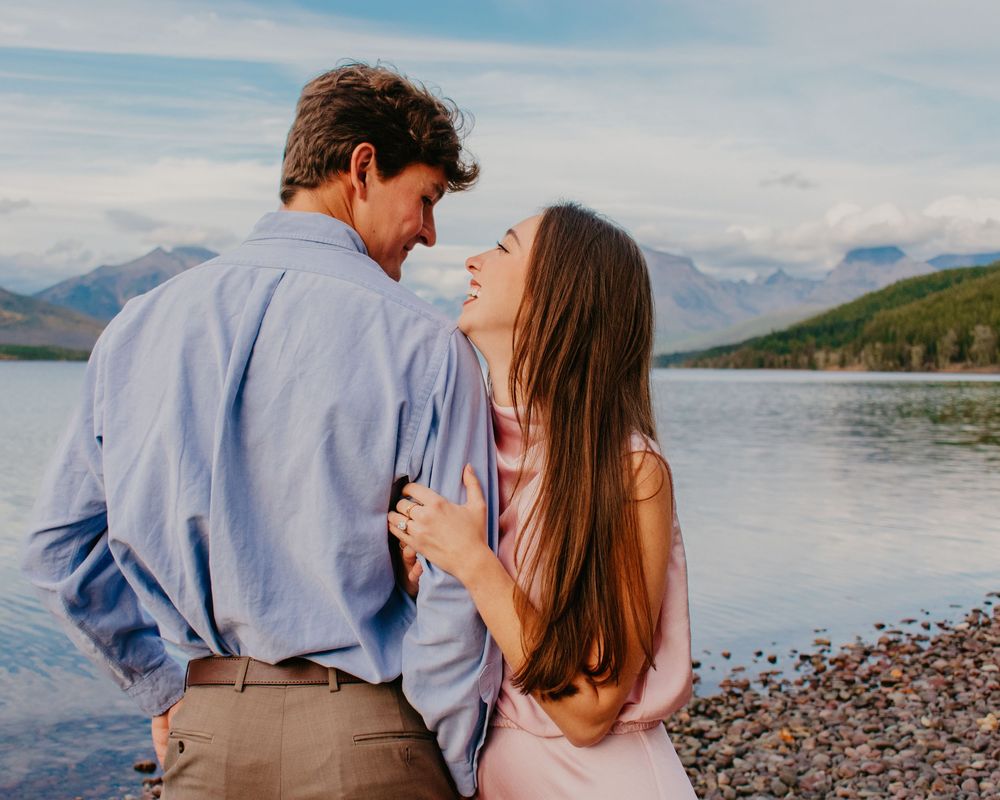 Romantic couple embraces on a rocky beach shore with mountains and lake in the background during sunset.