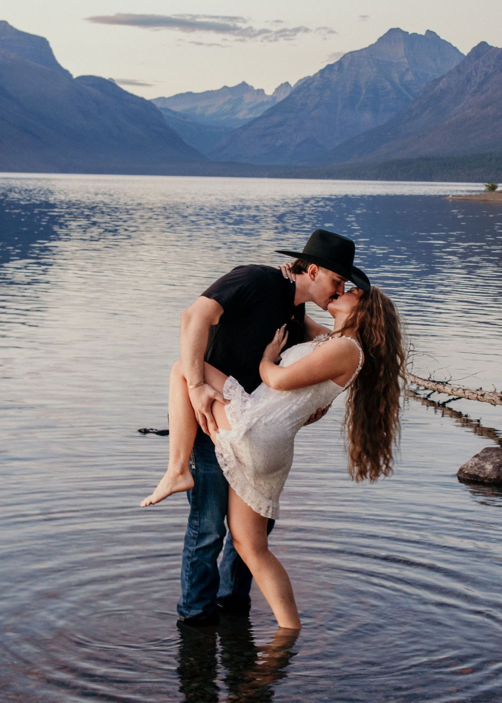 A couple in casual attire shares a romantic dip in a scenic mountain lake at sunset.