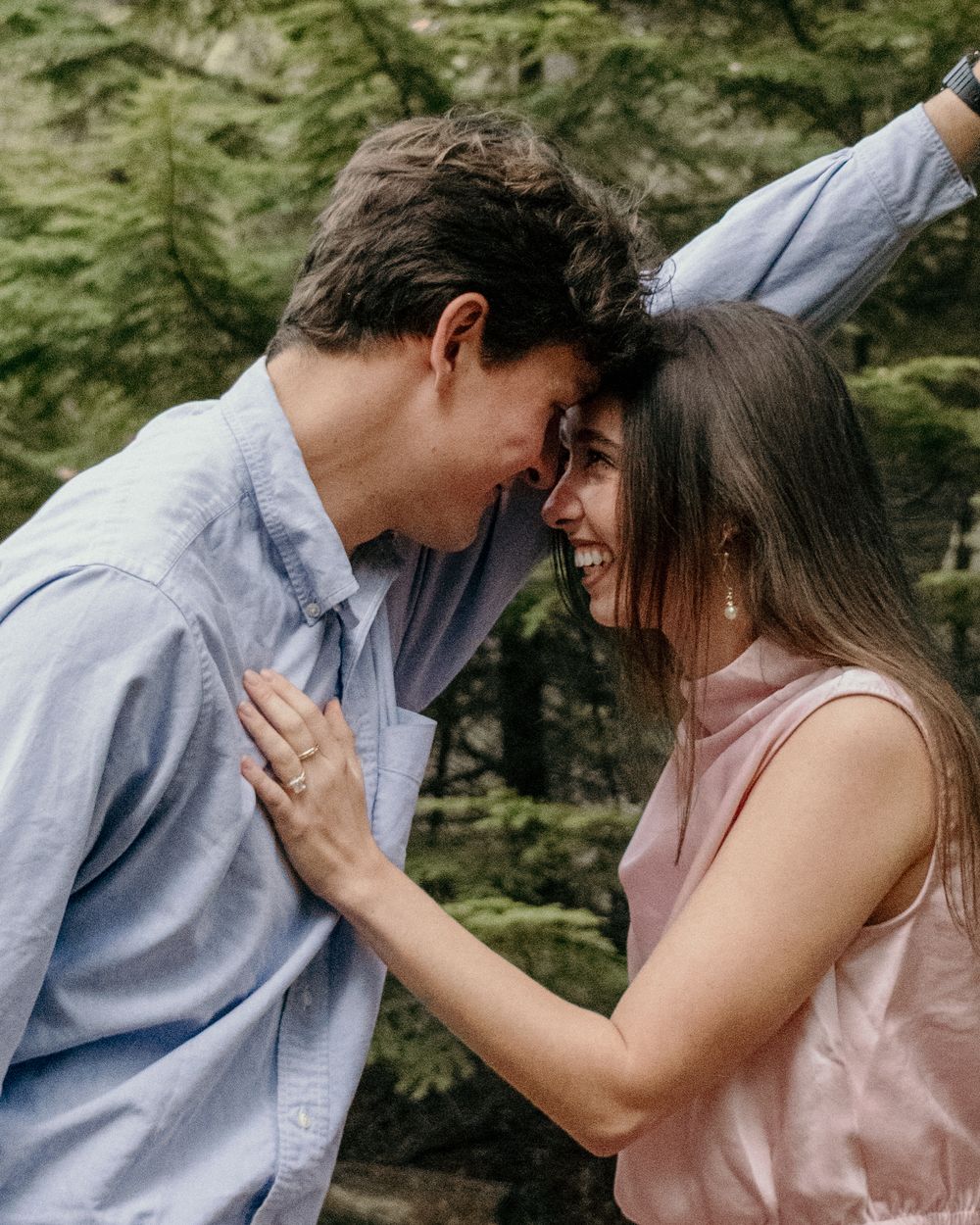 A romantic couple in casual attire share an intimate moment against a lush forest backdrop.
