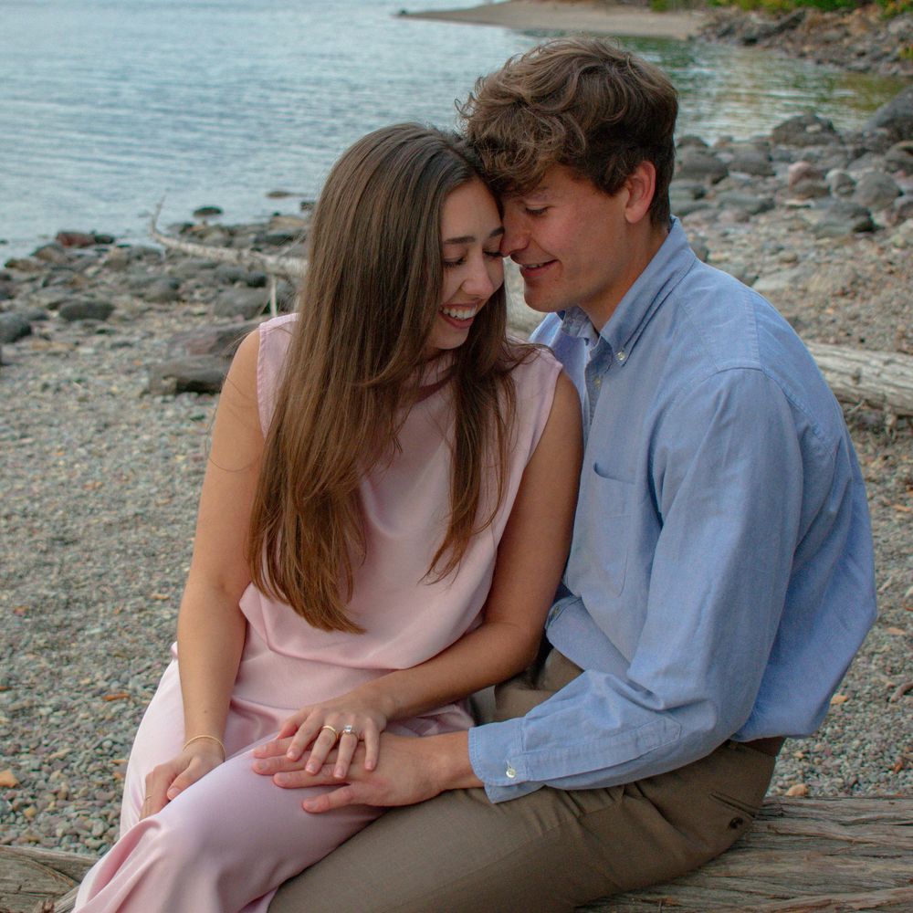 A romantic couple sits together on a rocky beach shore during sunset, sharing intimate moments by the water.