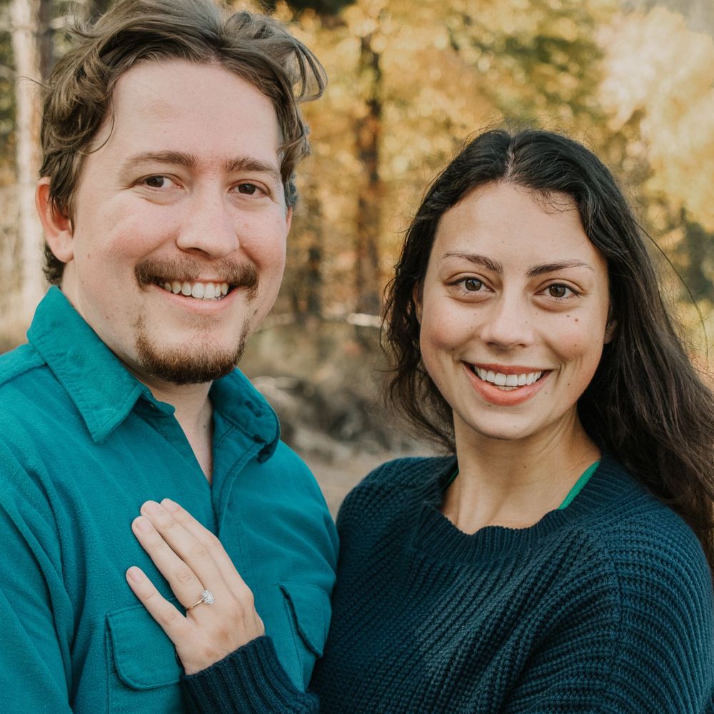 A couple stands close together outdoors in autumn, wearing a green shirt and black sweater while displaying an engagement ring.