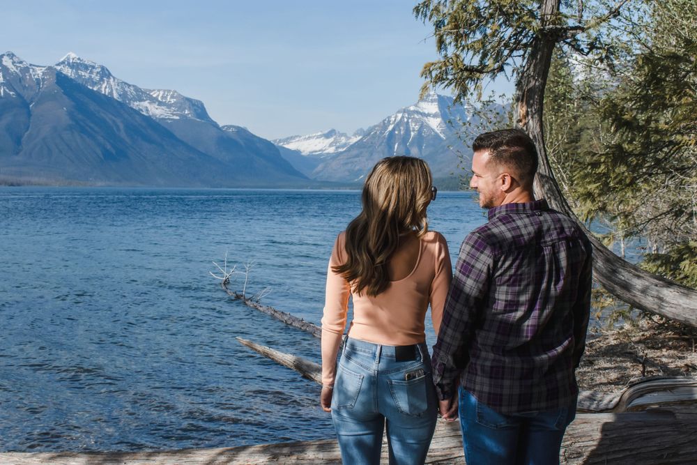 Couple walking along scenic mountain lake shoreline with snow-capped peaks in the background on a sunny day.