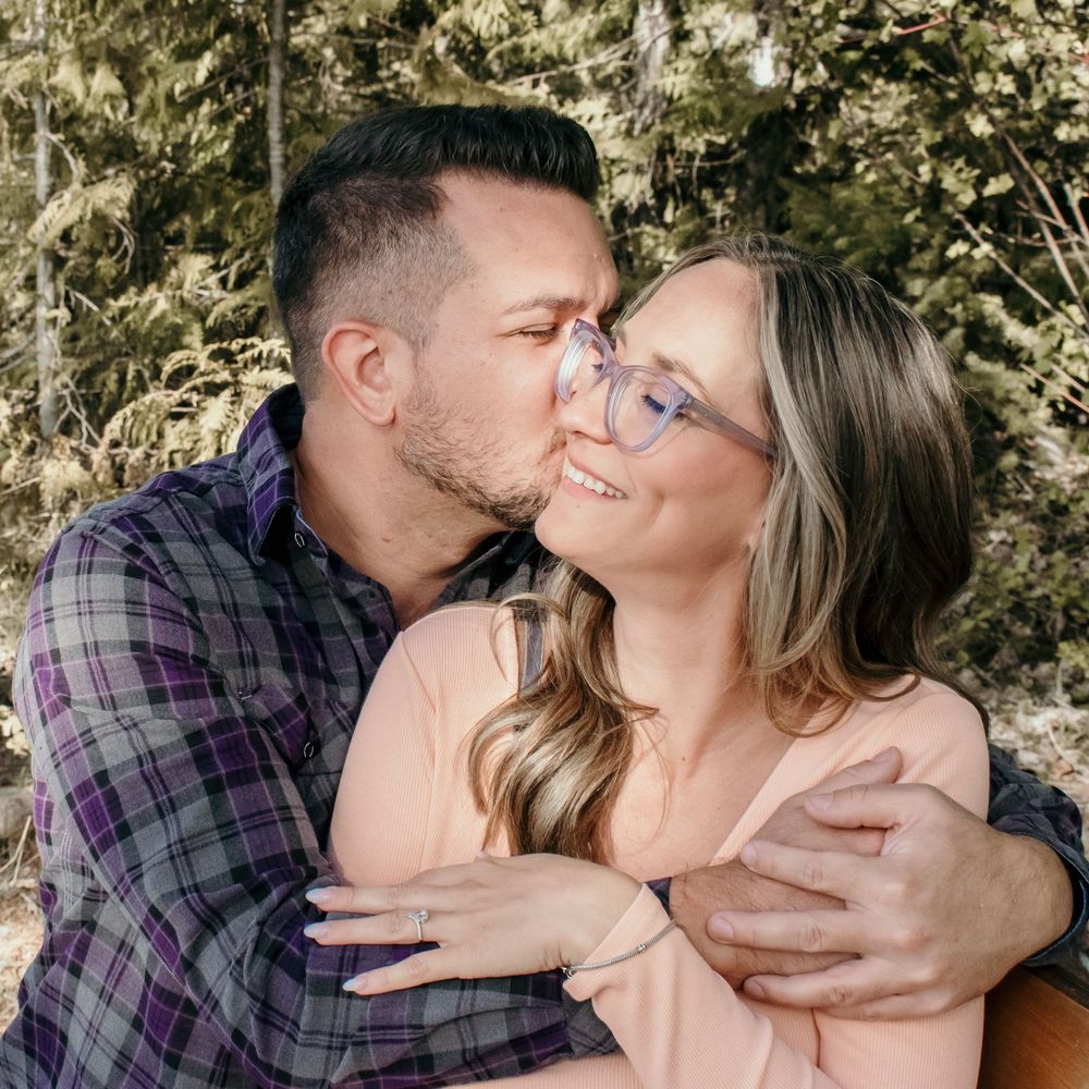 Couple in a romantic embrace sharing tender kisses against a natural outdoor backdrop with foliage.