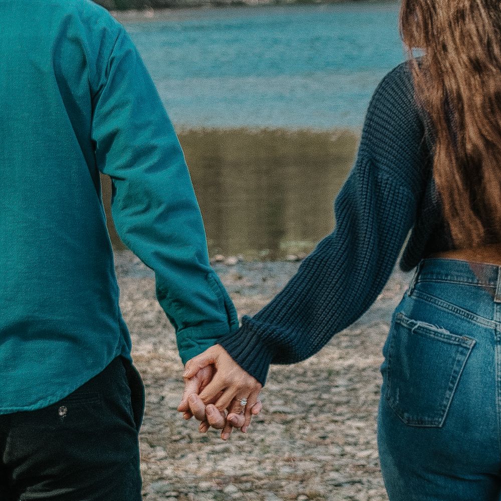 Two people holding hands while standing by a turquoise lake, wearing jeans and sweaters on a rocky shore.