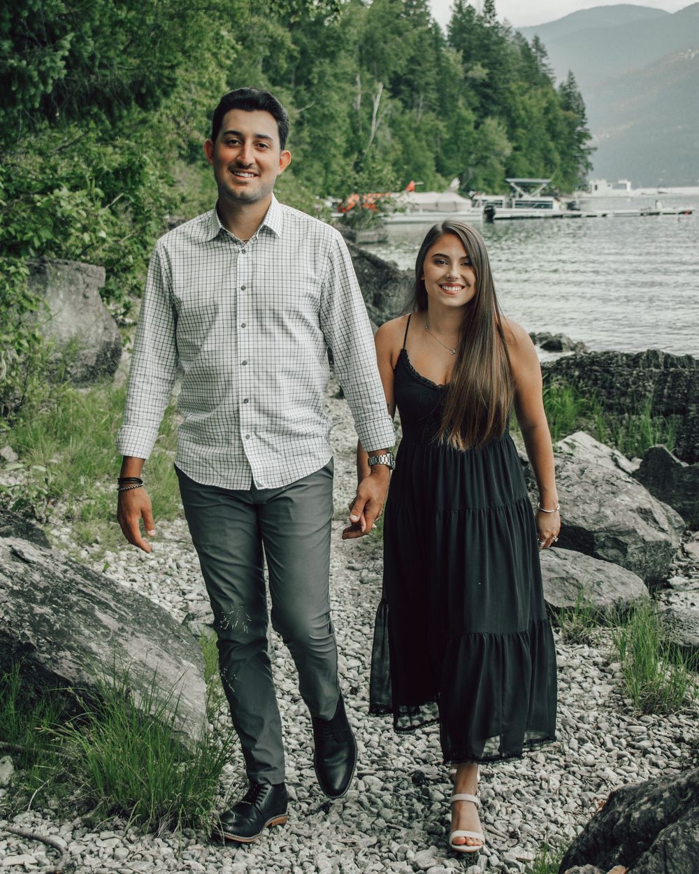 A couple stands together on a rocky lakeshore with mountains and trees in the background.