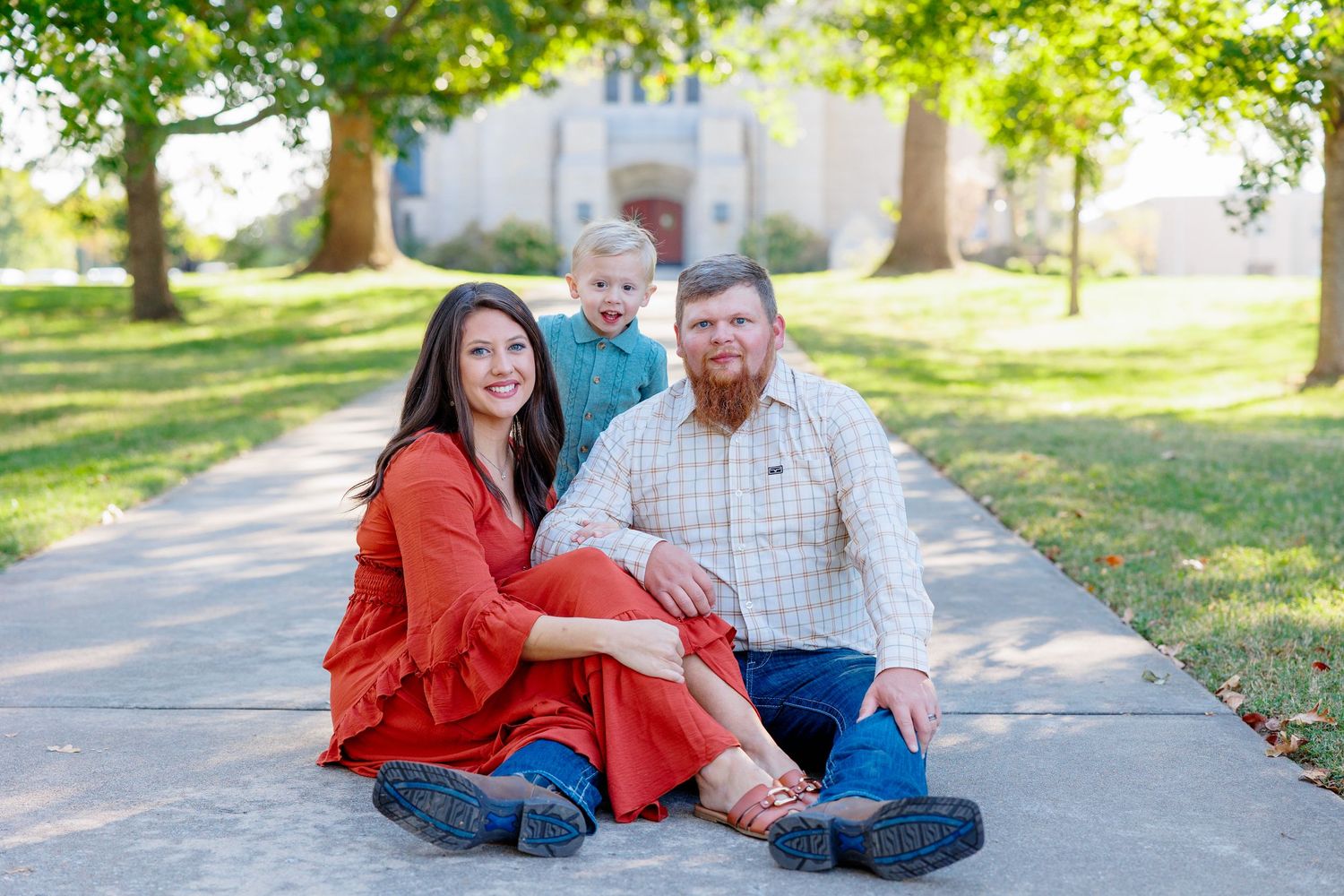 A couple in casual attire sits together on a paved path surrounded by green grass and trees in a park setting.