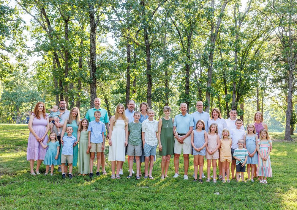A group of people wearing pastel colors stand together outdoors in a wooded area during a summer photo session in Branson, MO.