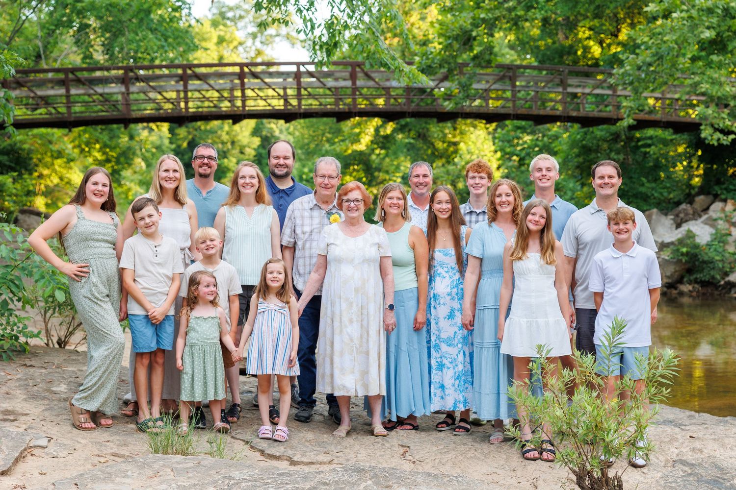 A family in coordinated blue and white outfits poses together outdoors near a rustic wooden bridge and creek in Branson, MO by Janie Loftin.