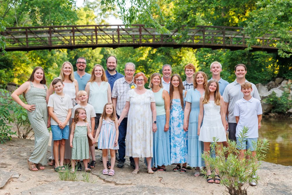 A family in coordinated blue and white outfits poses together outdoors near a rustic wooden bridge and creek in Branson, MO by Janie Loftin.