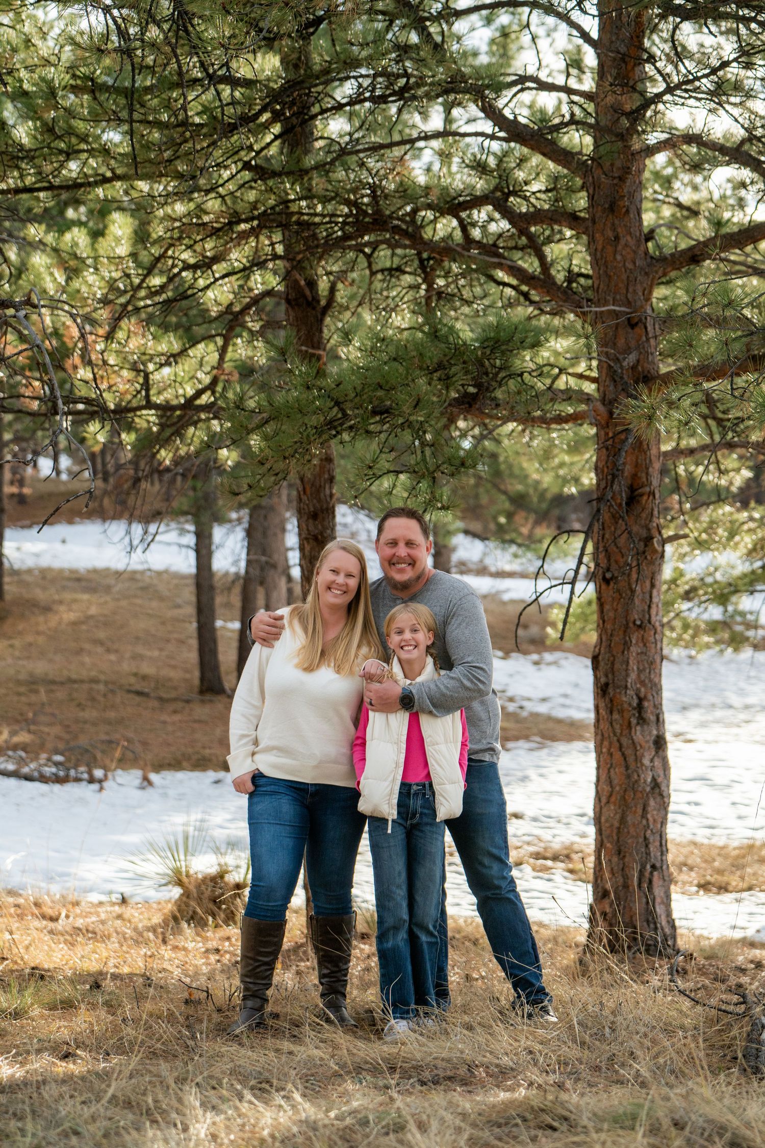 A family stands together among tall pine trees in a winter landscape with patches of snow on the ground.