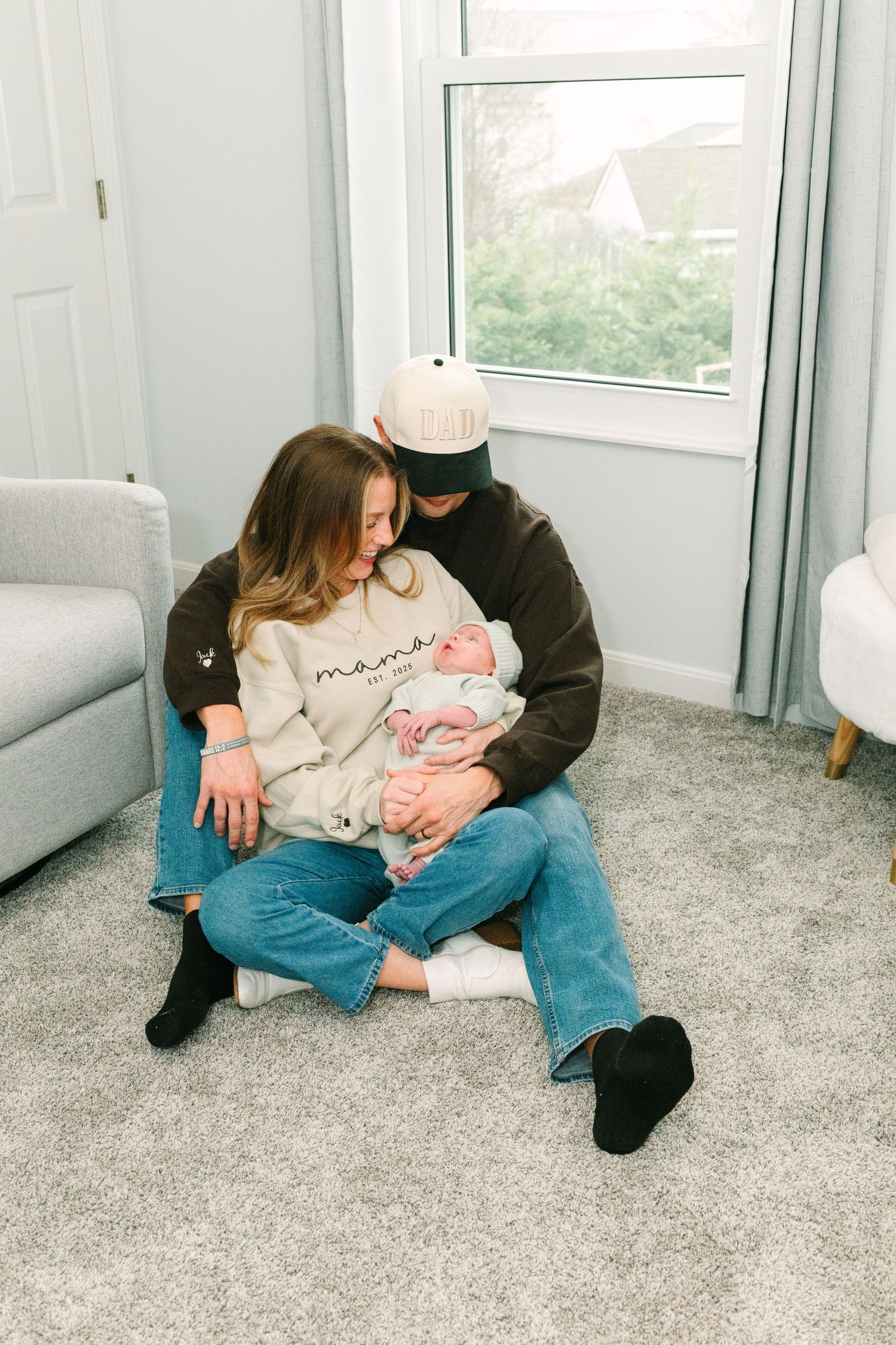 Expectant parents stand beside a white crib in a nursery with sage green accent wall.